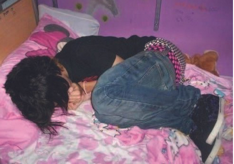 A person is curled up on a bed with pink cartoon-patterned bedding, wearing a black shirt, blue jeans, and a checkered accessory. The room features a purple wall, creating a playful and cozy atmosphere.