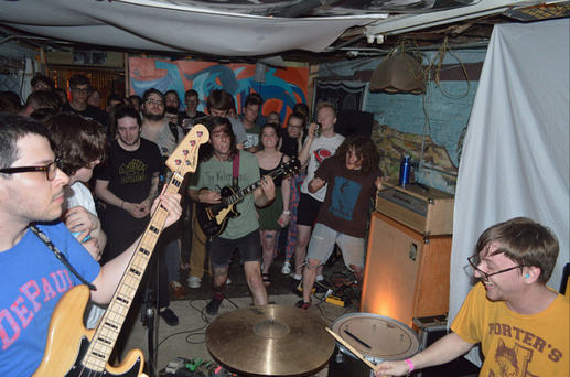 A DIY basement show featuring a punk or emo band performing in an intimate, crowded space. The bassist in the foreground wears glasses and a blue 'DePauw' shirt, playing a cream-colored bass. The drummer, in a yellow 'Porter's' shirt, is mid-motion with drumsticks in hand. The guitarist, wearing a green shirt, plays energetically while the audience, standing close, reacts with excitement. The setting includes exposed pipes, a low ceiling, and a mix of posters and makeshift decorations on the walls, adding to the underground venue’s raw atmosphere.