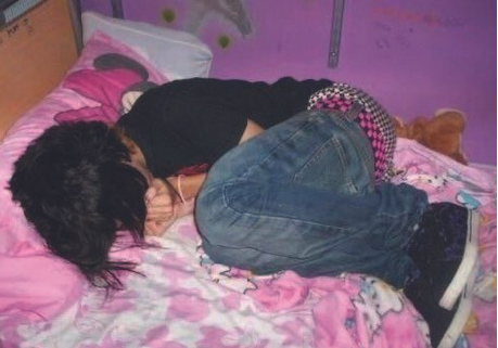 A person with dark, shaggy hair is curled up in a fetal position on a bed covered in pink, cartoon-themed bedding. They are wearing a black t-shirt, blue jeans, a checkered studded belt, and black sneakers with white soles. The room has purple walls decorated with plush toys and posters, evoking an early 2000s emo or scene aesthetic.