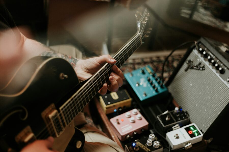 Close-up of a tattooed musician playing a black electric guitar, surrounded by an array of colorful guitar effect pedals and a vintage-style amplifier. The scene captures a warm, intimate atmosphere of music creation.